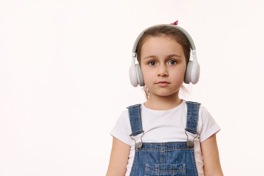 Adorable smart little child, cute baby girl wearing wireless headphones, looking at camera, isolated over white background. Studio shot with free space for your advertising and promotional text.