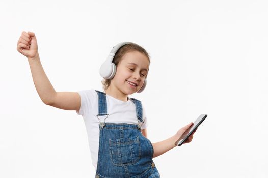 Lovely child, joyful baby girl in blue denim overall, holding smartphone and dancing while listening to the music on stereo headphones, isolated over white background with copy ad space for text