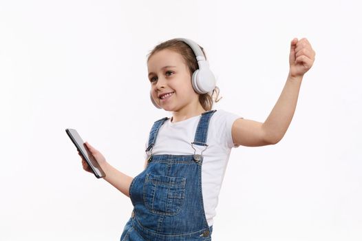 Joyful Caucasian child, a cute little girl wearing blue denim overalls, with smartphone in her hands, dances and listens to the music on her headphones, isolated over white background with copy space