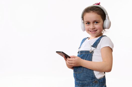 Isolated portrait on white background of a Caucasian smart little child, adorable baby girl in denim overalls, wearing wireless headphones, holding mobile phone and cutely smiles looking at camera