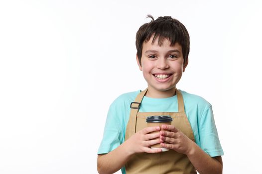 Multi-ethnic handsome preteen boy, wearing turquoise t-shirt and beige chef's apron holding an eco paper cup of takeaway coffee, smiling toothy smile looking at camera, isolated over white background