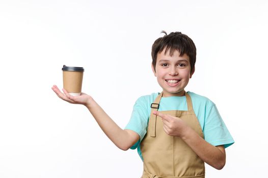 Lovely child boy wearing beige chef's apron, smiling a toothy smile looking at camera, pointing with finger at an eco-friendly cardboard mug of takeaway coffee, isolated white background. Copy space