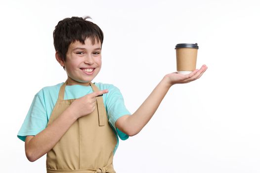 Multi-ethnic teenage boy wearing chef's apron, pointing with finger at a paper cup of takeaway coffee on his hands, isolated on white. Free space for your advertising. Food and drink consumerism.