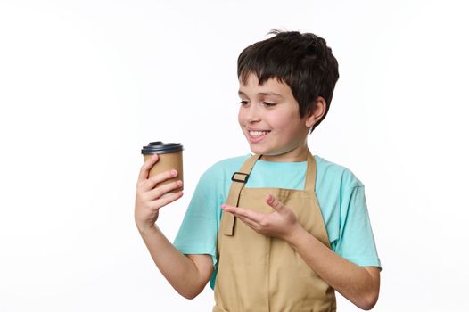 Awesome cheerful teenage boy - little barista, bartender in chef apron, selling a takeaway hot drink in ecological-friendly cardboard mug, isolated over white background. Beverage to go. Coffee house