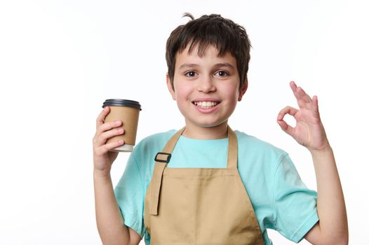 Isolated portrait on white background of a happy teenage boy bartender showing OK sign, selling a takeaway coffee beverage drink in eco paper cup. Hot beverage to go. Copy space for your advertising