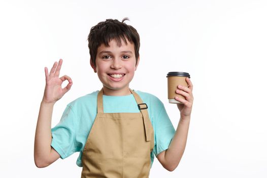 Amazing teenage boy - little barista bartender in chef apron, showing OK sign while selling hot takeaway drink in paper mug, smiling looking at camera, isolated on white background with free ad space