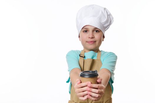 Multi-ethnic preteen boy bartender, wearing beige apron and white chef's hat, handing out on camera a takeaway hot tea coffee drink in cardboard mug. Beverage to go. Free space for promotional text.