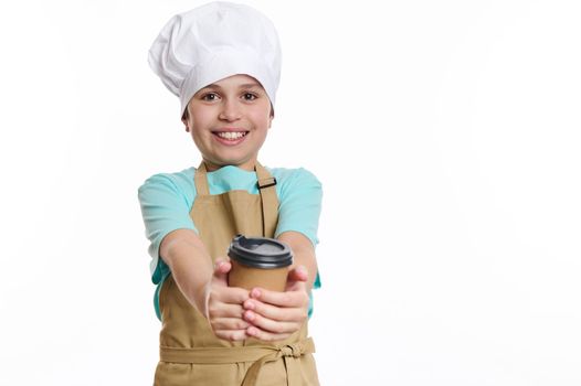 Isolated portrait on white background of a lovely teenage boy, bartender barista, wearing chef's hat and apron, holding selling making a hot tea coffee drink in eco paper cup to go, looking at camera