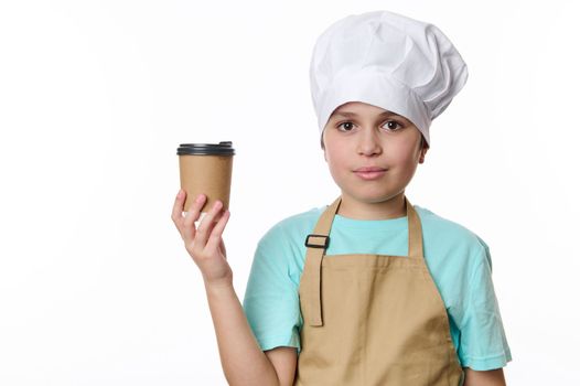 Adorable multi-ethnic teenage boy - barista barman bartender wearing white chef's hat and beige apron, looking at camera while holding a takeaway hot drink in cardboard cup to go, isolated on white