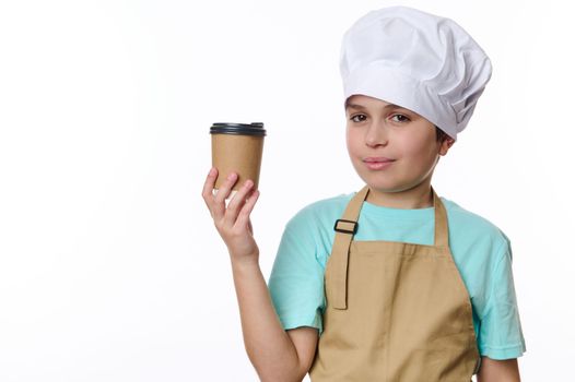Handsome Middle-Eastern teenage boy in chef's hat and beige apron, selling a takeaway hot beverage coffee tea in eco paper cup to go, cutely smiling looking at camera, isolated over white background