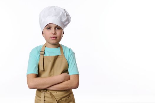 Confident portrait of a handsome teenage boy, dressed as a cook or baker confectioner in chef's hat and beige apron, isolated on white background with free space for your advertising text.