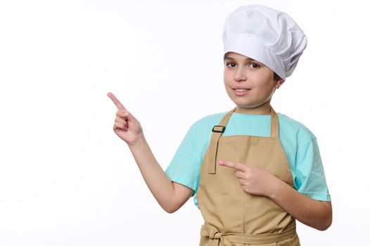 Adorable teenage boy dressed as a chef pastry or baker confectioner, wearing a white hat and beige apron, looks at camera and points at free space for promotional text, isolated over white background