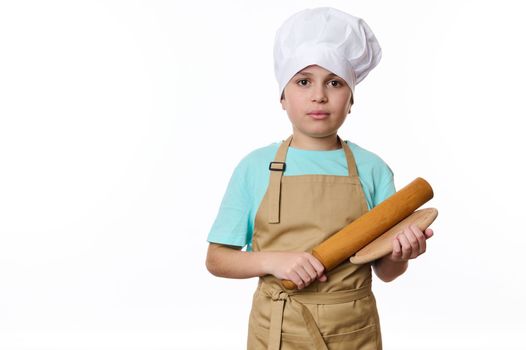 Isolated portrait on white background of a handsome teenage boy, wearing a white chef's hat and beige apron, a cook, baker confectioner posing with wooden board and rolling pin. Free advertising space