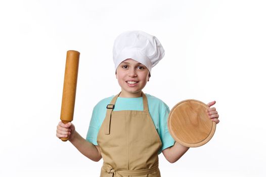 Adorable Caucasian teenage boy - baker wearing chef uniform, smiles a cheerful toothy smile, posing to camera with a wooden cutting board and rolling pin in his hands, isolated over white background