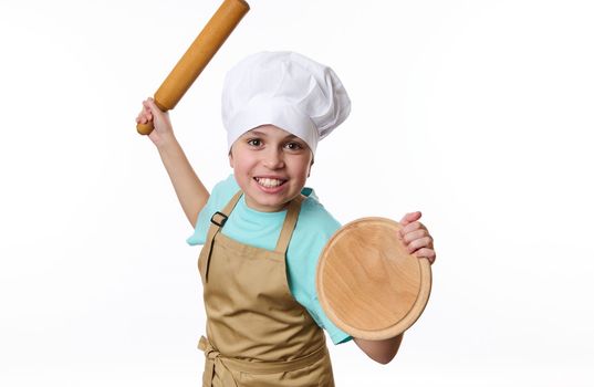 Isolated portrait on white background of a playful teenage boy, baker wearing chef's hat and apron, fighting with a wooden board and rolling pin in his hands, looking at camera. Free advertising space