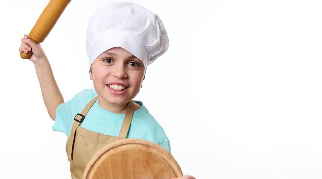 Close-up of a smiling mischievous boy - baker confectioner wearing chef's hat and apron, fighting with wooden board and rolling pin in his hands, isolated on white background. Bakery advertising shot.
