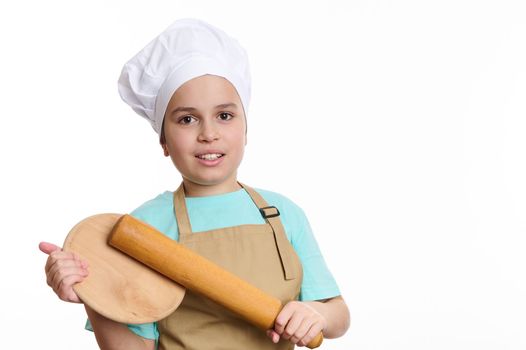 Multi-ethnic teenage boy - baker wearing chef uniform, smiles a cheerful toothy smile, posing to camera with a wooden cutting board and rolling pin in his hands, isolated over white background