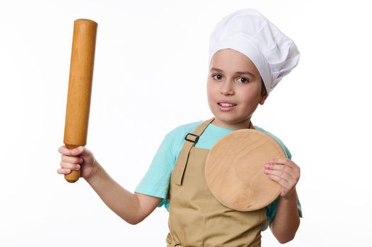 Adorable Caucasian child boy, wearing a chef's hat and beige apron, posing with a rolling pin and wooden cutting board, isolated on white background. Baker Confectioner. Chef pastry. Cook profession