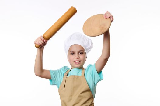 Multi-ethnic overjoyed happy pre-adolescent child, little baker confectioner, cook in chef apron and hat, holds a wooden board and rolling pin, looks at camera, isolated white background. Free space