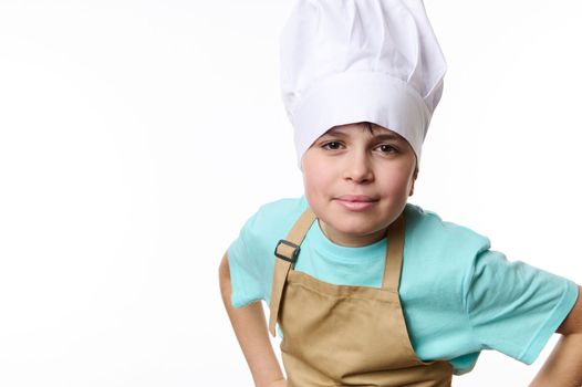 Isolated portrait on white background: teenager boy - chef pastry, cook, baker confectioner wearing a chef hat and beige apron, holding hands on belt, smiling looking at camera. Free advertising space