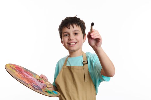 Artistic teenage boy wearing beige apron, holding a paintbrush and a color palette, paints on imaginary canvas, looking at camera, isolated on white background. Kids entertainment and art development