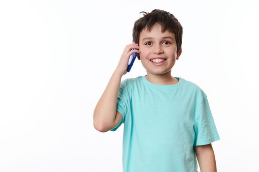 Overjoyed excited dark-haired Caucasian boy, wearing turquoise t-shirt, cutely smiling a cheerful toothy smile looking at camera while talking on mobile phone, over white background with copy space