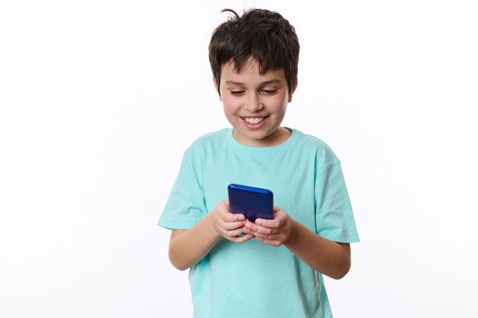 Isolated portrait on white background of a joyful happy teenage boy, wearing turquoise t-shirt, playing virtual online games on his smartphone. Digital gadget and entertainments for kids. Copy space