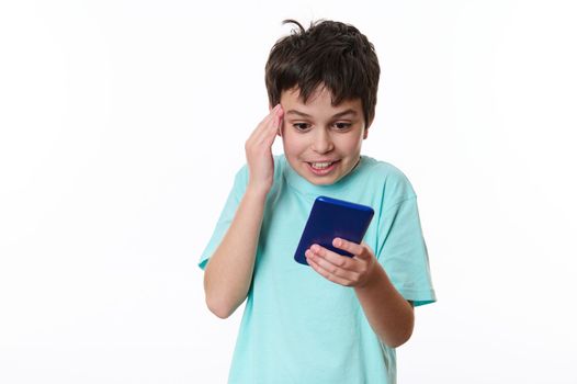 Surprised overjoyed multi-ethnic teenager, schoolboy wearing blue t-shirt, holding his hand on his temple, expressing astonishment while reading news on his smartphone, isolated over white background