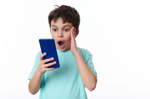 Shocked Caucasian teenage boy using mobile phone, reading news, expressing shock, surprise, stupefaction, astonishment, isolated over white background. Copy advertising space for promotional text