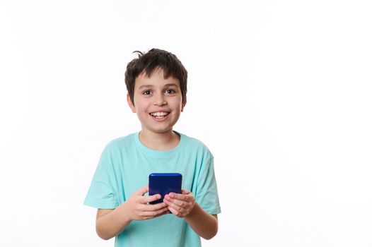 Waist-length portrait, isolated over white background of a smiling happy friendly teenage boy in blue turquoise t-shirt, using mobile cell phone, browsing surfing internet. People, lifestyle concept