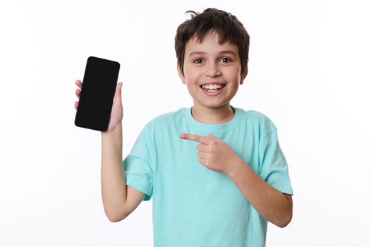 Wow awesome mobile app. Handsome Caucasian teenage boy points at smartphone with black blank screen with copy space for young advertising text, smiles looking at camera, posing over white background