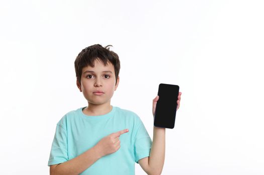 Child boy in blue t-shirt points at a smartphone with blank screen for your advertising or mobile application. Kids and digital gadgets. Studio shot of a teenager with cell phone on white background