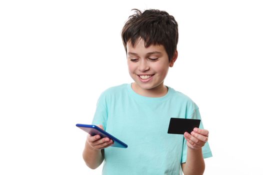 Isolated portrait on white background of a cheerful handsome smiling Caucasian teenage schoolboy, using mobile phone and credit card, makes cashless payment while shopping online, Internet banking