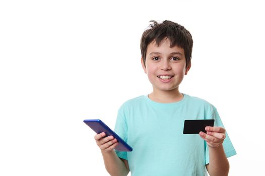 Adorable dark-haired Caucasian preteen boy, smart school kid in blue t-shirt with smartphone and plastic credit card, isolated on white background. Internet banking. Cashless payment. Online shopping