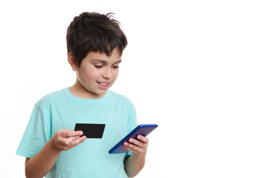 Close-up portrait of a smart teenage boy in a blue t-shirt, transfers data from a credit card to a mobile application on his smartphone, makes a cashless online payment, isolated on a white background