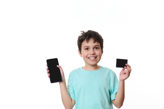 Adorable teenager boy smiles happily at camera, showing a smartphone and a black credit card with free space for advertising text, isolated on a white background. Internet banking. Mobile apps concept