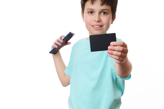 Selective focus on black credit card with blank space for your promotional text in the hands of a blurred teenage boy, smiling a cheerful toothy smile looking at camera, isolated over white background