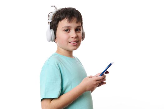 Isolated portrait on white background of a Caucasian smart teenage boy, wearing wireless headphones, using mobile phone, checking social media content, surfing the net, cutely smiles looking at camera