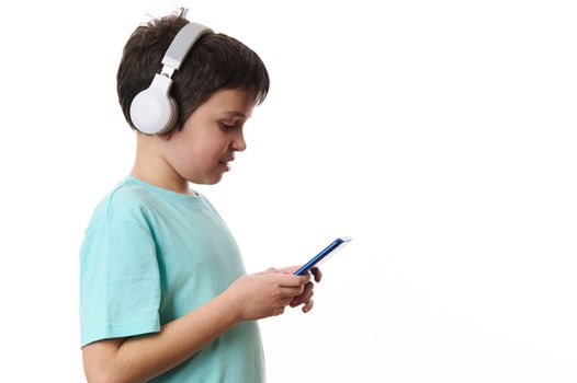 Side portrait of a concentrated teenage boy wearing wireless headphones, using mobile phone, checking social media content, surfing the Internet, browsing websites, isolated over white background.