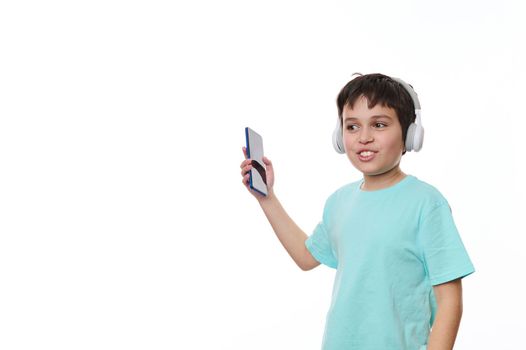 Lovely child, teenage boy wearing blue turquoise t-shirt, holding smartphone and dancing while listening to the music on stereo headphones, isolated over white background with copy ad space for text
