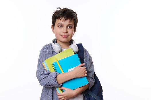 Confident handsome teen boy, elementary school student with headphones, looking at camera, posing with school supplies and backpack over white background. Back to school on new academic year. Ad space