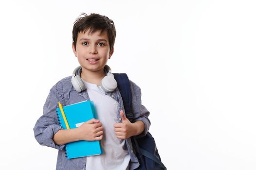 Caucasian adorable primary school student, smart teenage boy in casual wear, with wireless headphones, backpack and school supplies, looks at camera on white background. Copy ad space. Back to school