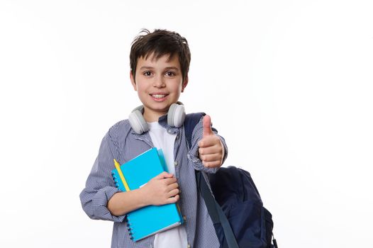 Caucasian smart primary school student, teenage boy in casual wear, with wireless headphones, backpack and school supplies, thumbs up looking at camera, white background. Copy ad space. Back to school