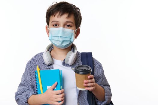 Close-up portrait of a handsome schoolboy in casual clothing, wearing protective medical mask, holding out a disposable paper cup with hot drink, looking at camera, isolated on white background
