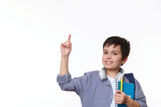 Middle-Eastern teenage school boy in casual wear, expressing positive emotions, pointing at copy advertising space, smiling at camera, posing with school supplies and backpack over white background