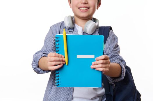 Focus on a blue copybook with yellow pen in hands of a cheerful teenage school boy, wearing casual wear, smiling a toothy smile, isolated on white background, copy space. School and education concept