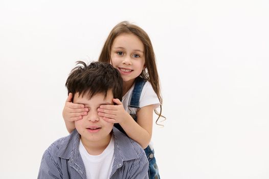 Adorable little child girl closes her brother's eyes with her hands, gently hugs him from the back, sweetly smiles looking at camera, isolated over white background. Friendships. Family relationships