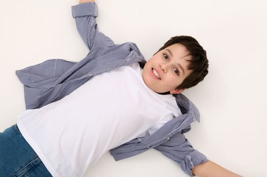 View from above of Caucasian adorable teen boy in blue jeans, checkered shirt and white t-shirt, looking at camera, lying down on a white background. Copy advertising space. Portrait of beautiful kids