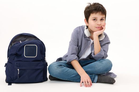 Lovely Caucasian boy teenager, elementary school student wearing casual denim, with blue backpack, confidently looking at camera, isolated on white background. Copy advertising space. Back to school