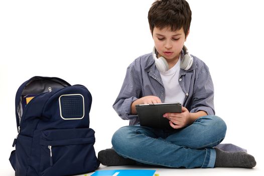 Caucasian handsome teenage schoolboy with wireless headphones, in casual wear, using digital tablet, studying online, doing homework, isolated on white background. Back to school. Knowledge Education